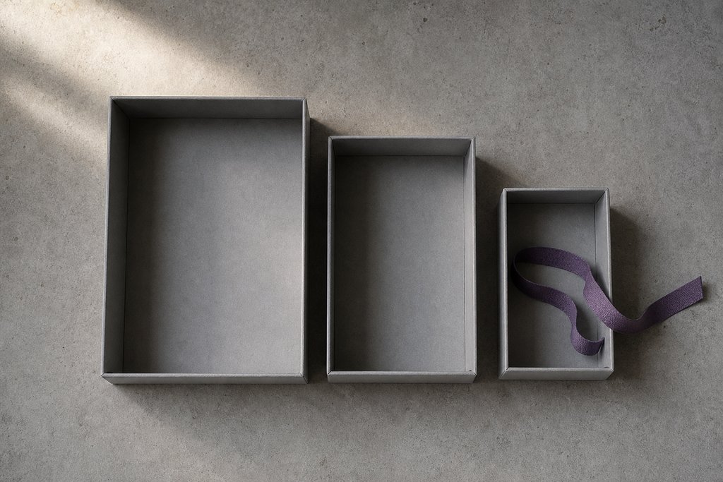 Drei graue Archivkartons abnehmender Größe nebeneinander, alle leer und unbeschriftet, ein violettes Bindeband über dem kleinsten