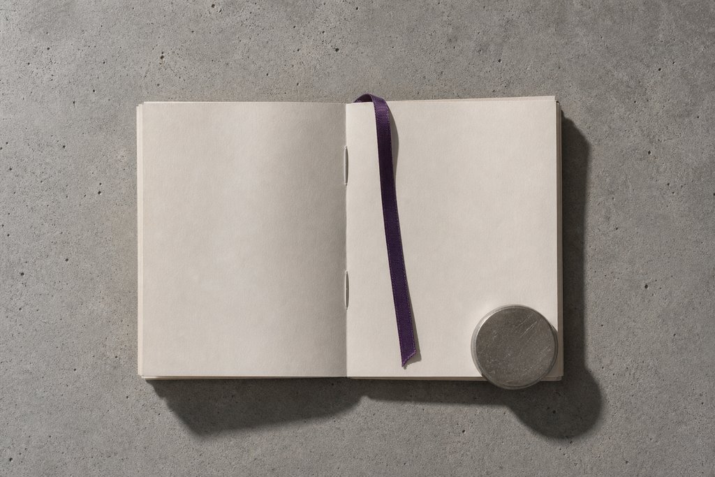 Aufgeschlagener Stapel leerer heller Seiten, ein violettes Band markiert eine Seite, ein Stahlgewicht hält die Ecke