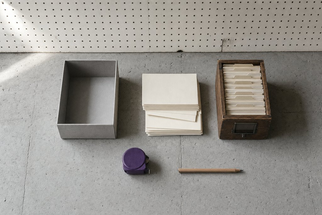 Offener grauer Archivkarton mit leeren Karteikarten, eine hölzerne Karteischublade und ein violettes Maßband auf Sichtbeton